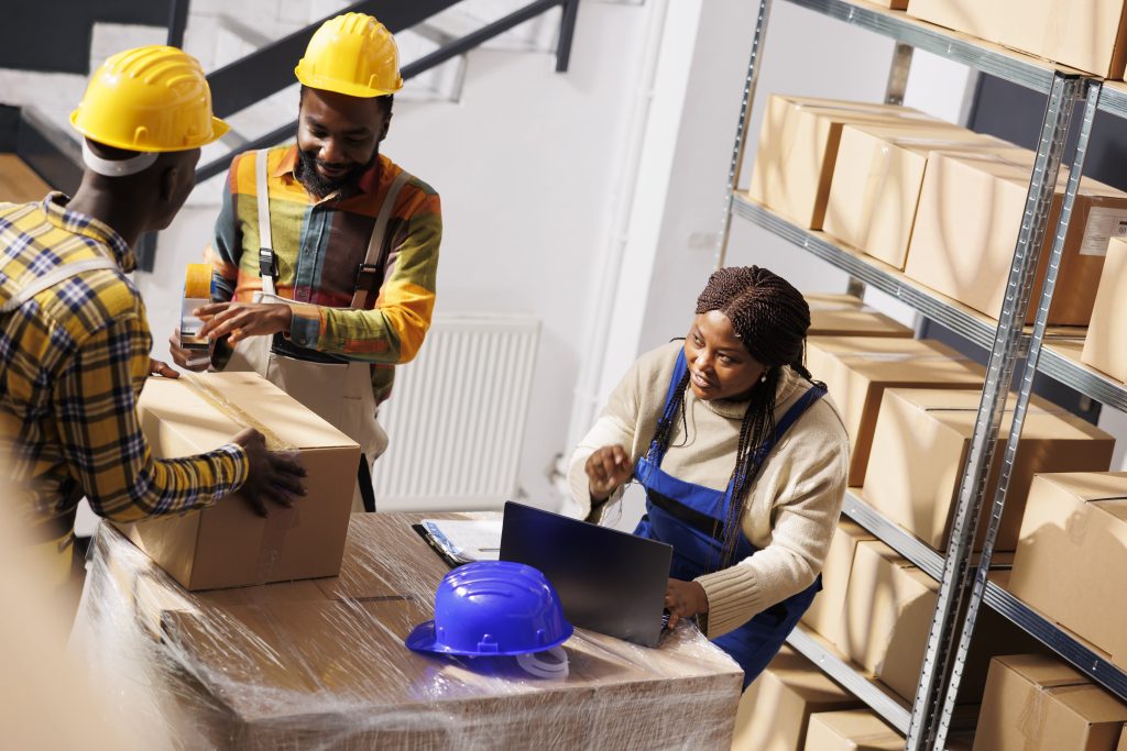 warehouse manager checking orders list coordinating parcels packing process african american woman supervisor with laptop controlling freight cardboard boxes sealing dispatching min